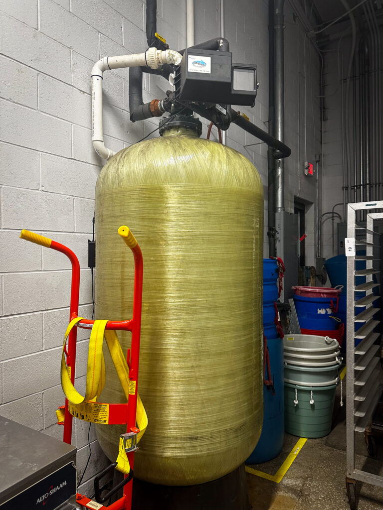 A large cylindrical tank with pipes and a black box at the top. It is a charcoal filtration system. 