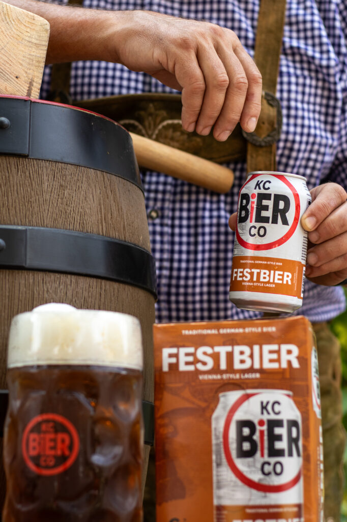 A man can be seen from the shoulders down in a blue checkered shirt and lederhosen. He is resting one elbow on a small barrel, and is holding a can of KC Bier Co Festbier on top of a 6-pack of KC Bier Co Festbier
