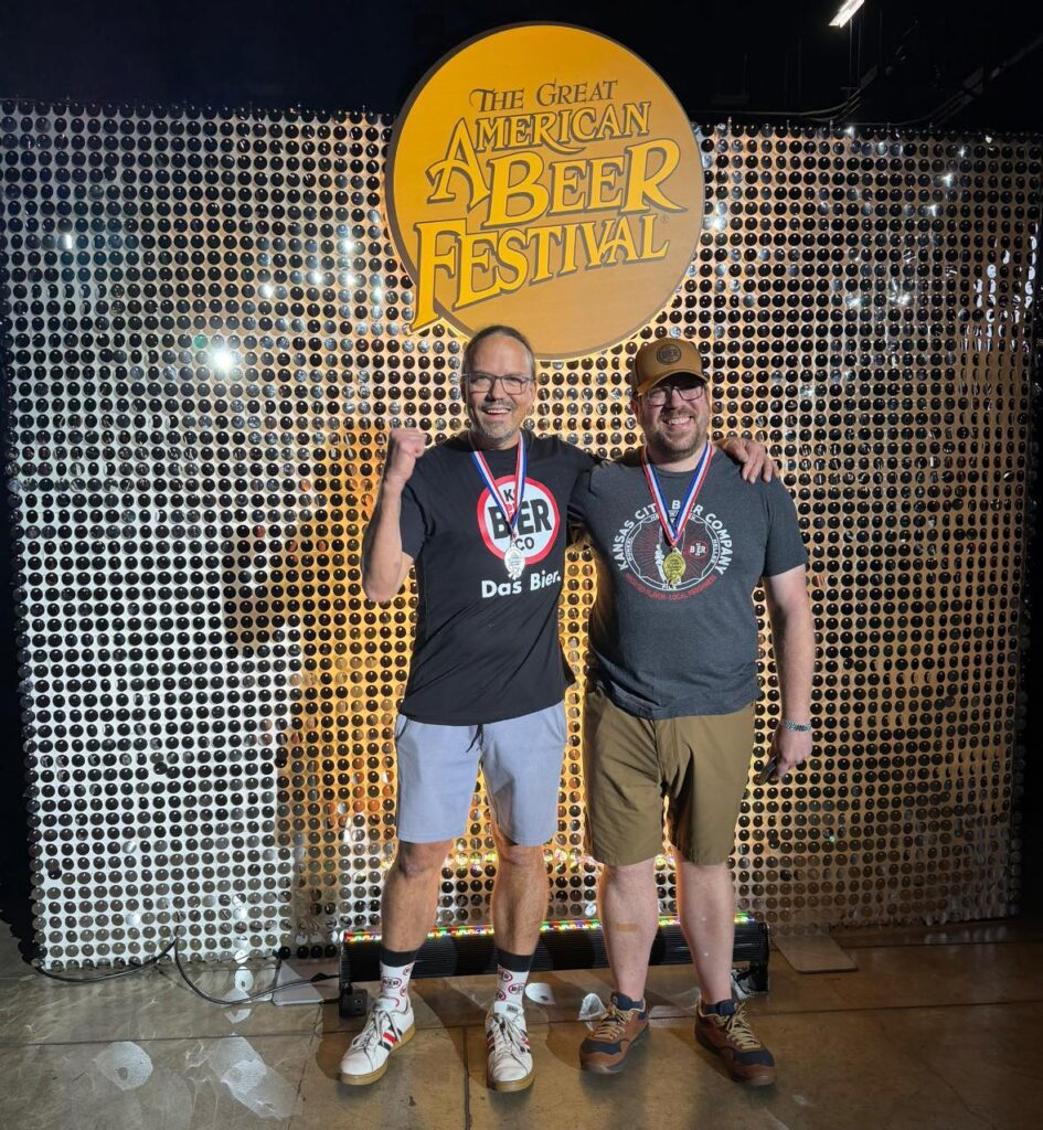 Two men stand on stage against a shiny silver backdrop and a Logo that says Great American Beer Festival. They are happy, with medals around their necks. 