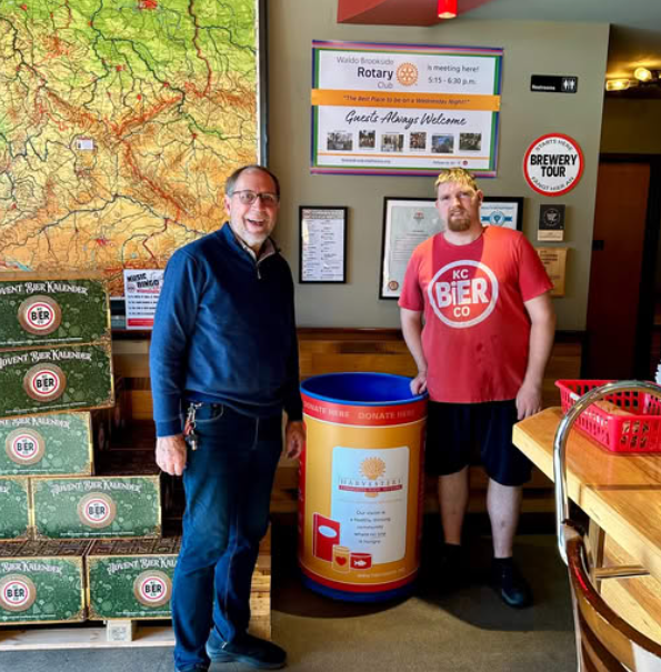 Two men stand beside a blue barrel. The barrel has Harvesters Kansas CIty logos and information on it. The men are smiling