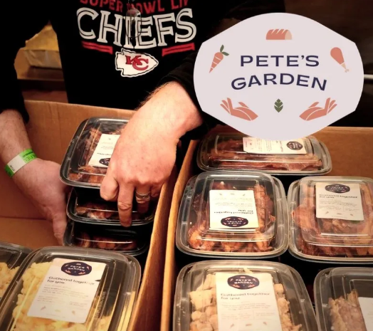 Cardboard boxes are filled with plastic meal containers with clear lids. The stickers on containers say Pete's Garden. A man in a Kansas City Chiefs long-sleeve shirt loads more containers into the box. 
