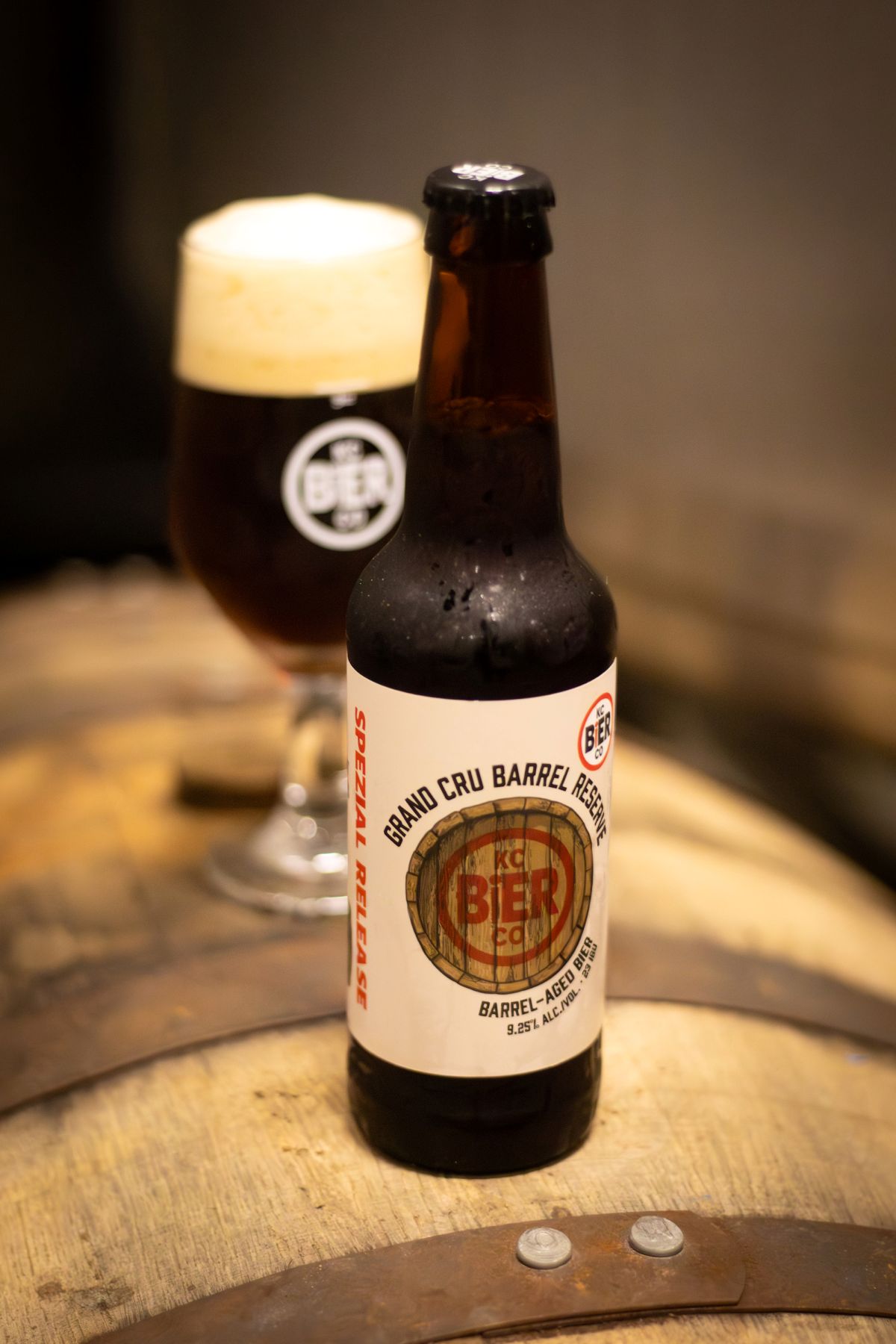 A photo of a bottle of beer sitting on top of a wooden whiskey barrel. The bottle says "Grand Cru Barrel Reserve" and features artwork of an illustrated barrel. Behind the bottle is an out of focus stemmed glass that has dark brown beer in it.
