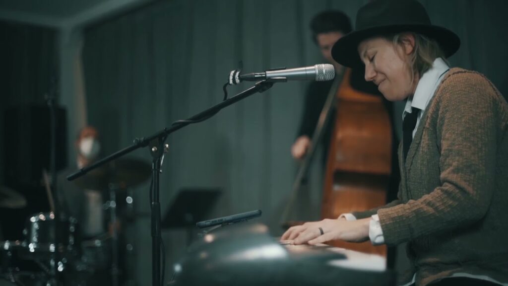 A photo that shows a smiling woman in a top hat playing an electric keyboard in front of a microphone. In the background, a standing bass can be seen. 