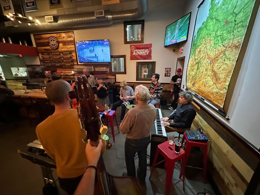 The image shows 5 musicians from a fish-eye view, with the photo-taker holding a tambourine. There is a keyboard, xylophone, guitar, and more. Someone is standing back and taking a photo of the musicians. 